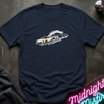 A navy blue short-sleeve crewneck t-shirt featuring a graphic print of a white sports car leading a line of other cars, laid flat on a dark textured surface with a neon sign and dark pants partially visible.