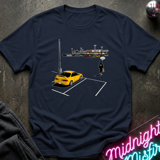 A navy blue short-sleeve t-shirt with a graphic print of a yellow sports car in a parking spot and a small figure, laid flat on a dark textured surface with a neon sign and sweatpants partially visible.
