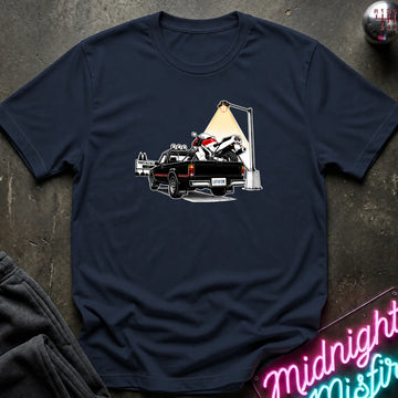 A navy blue short-sleeve crewneck t-shirt featuring a graphic print of a black pickup truck carrying a white and red motorcycle under a street lamp, laid flat on a dark textured surface.