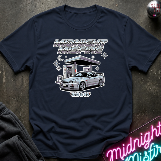 A navy blue graphic t-shirt laid flat on a dark textured surface. The shirt features a white and silver graphic of a sports car at a gas station with the text 'MIDNIGHT MISFIRE' and 'GAS GAS'.