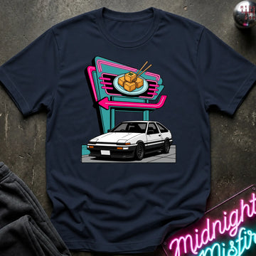 A navy blue short-sleeve crewneck t-shirt featuring a retro-style graphic of a white sports car parked in front of a neon sign. The shirt is displayed flat on a dark textured surface with a dark sweatshirt and a neon sign partially visible as props.