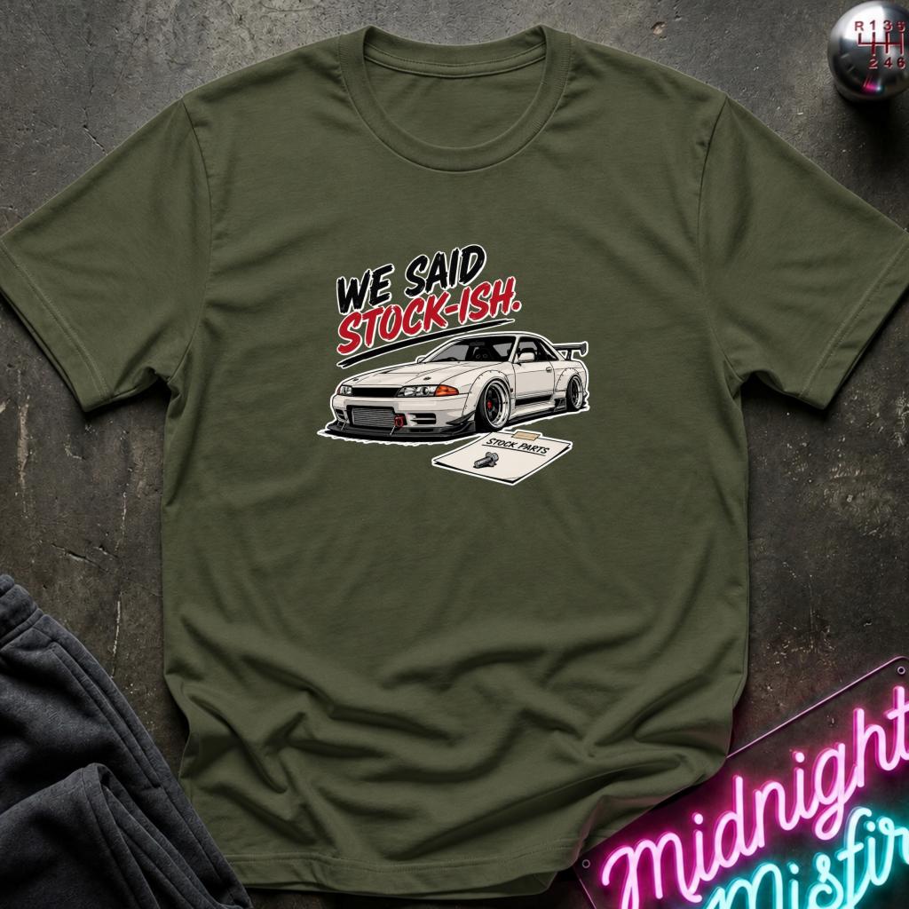 An olive green crewneck t-shirt featuring a graphic of a white sports car and the text 'WE SAID STOCK-ISH!' in black and red, laid flat on a dark textured surface.