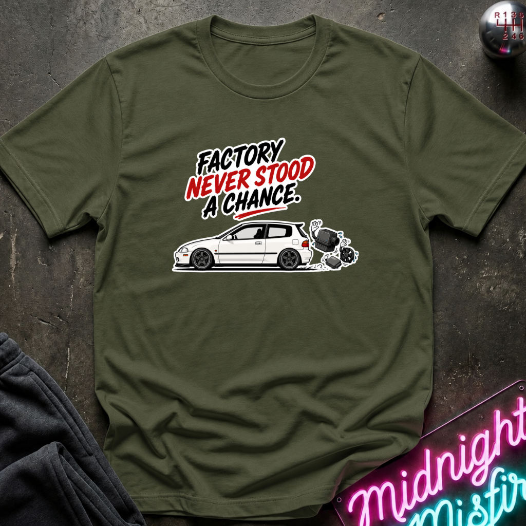An olive green short-sleeve t-shirt with a graphic print on the front. The graphic features the text 'FACTORY NEVER STOOD A CHANCE' in black and red lettering above a white car and a small dog illustration.