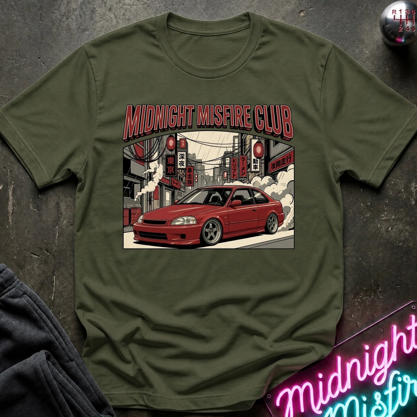 An olive green crewneck t-shirt featuring a graphic print of a red coupe car and the text 'MIDNIGHT MISFIRE CLUB' in red, styled flat on a dark textured background with a neon sign and dark sweatpants partially visible.
