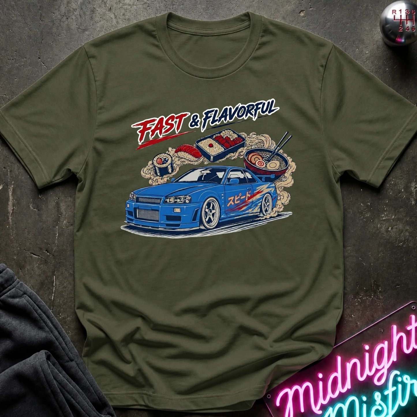 An olive green short-sleeve t-shirt with a graphic print of a blue sports car, sushi, ramen, and the text 'FAST & FLAVORFUL' in red and white. The shirt is displayed flat on a dark textured surface with a neon sign and dark fabric partially visible in the corners.