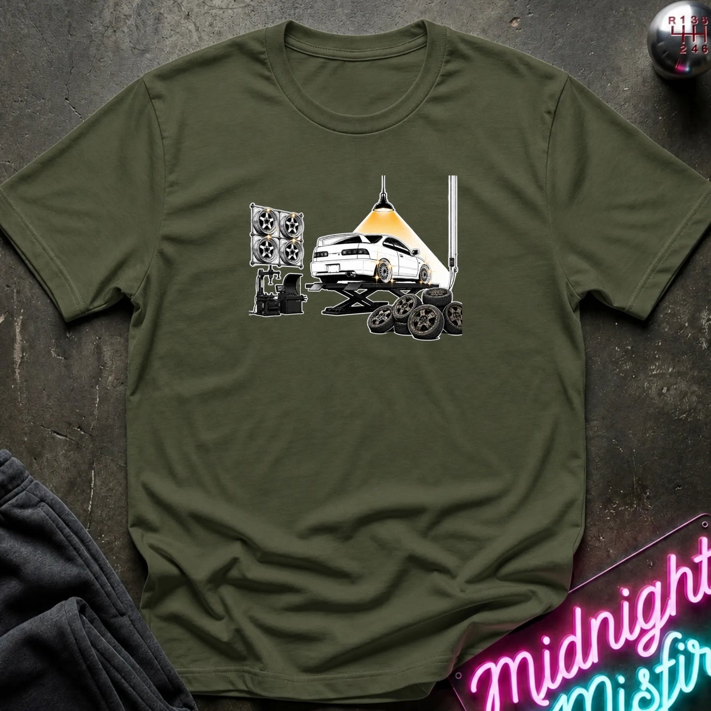 An olive green short-sleeve crewneck t-shirt laid flat on a dark textured surface. The shirt features a graphic print of a white car on a hydraulic lift with new wheels.