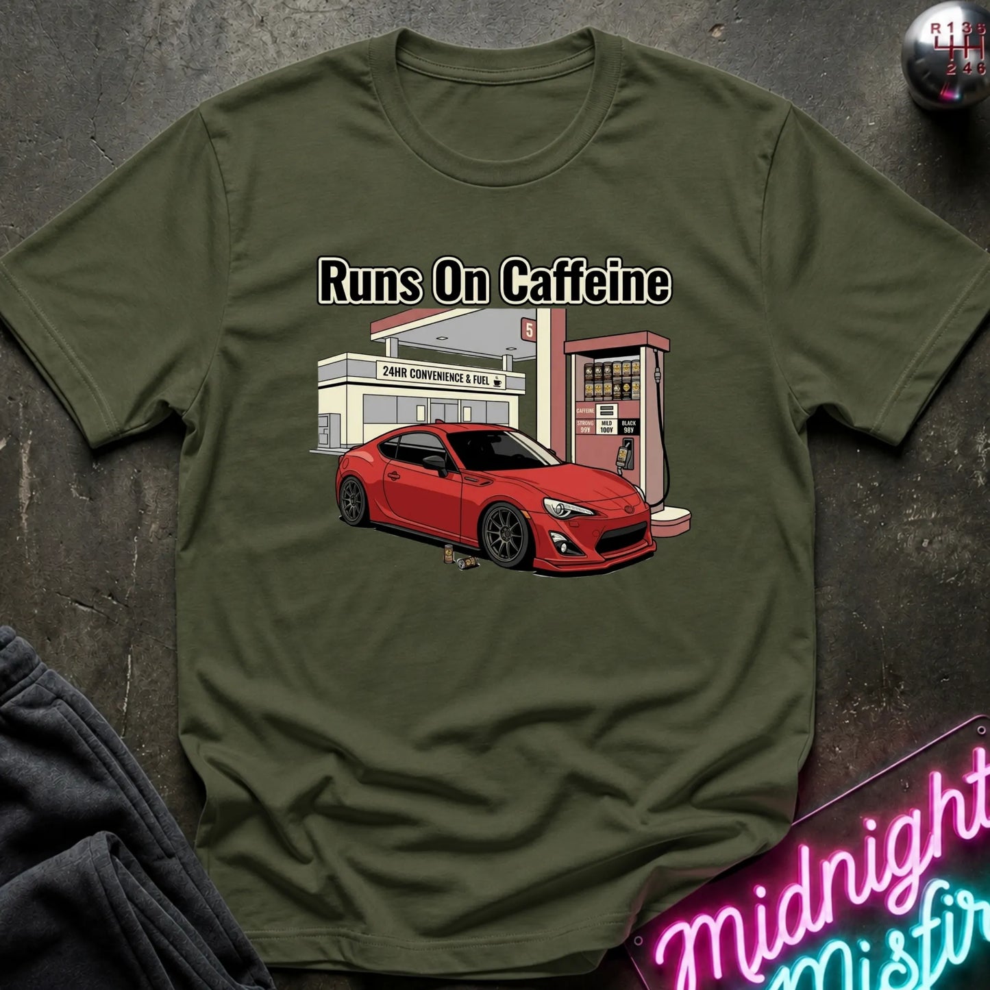 An olive green graphic t-shirt laid flat on a dark textured surface. The shirt features a cartoon illustration of a red sports car parked in front of a convenience store, with the text 'Runs On Caffeine' printed above the graphic.
