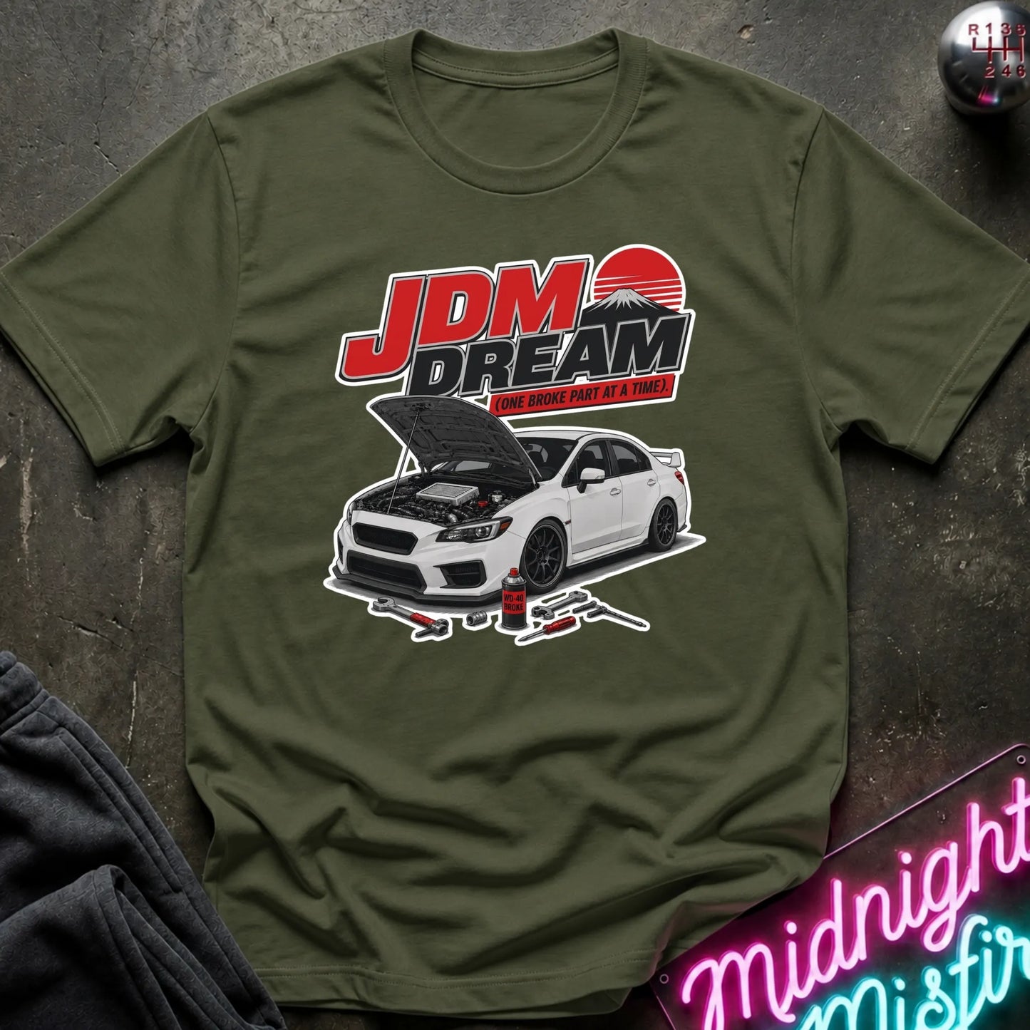 An olive green graphic t-shirt laid flat on a dark textured surface. The shirt features a graphic of a white car with its hood open under the red and black 'JDM DREAM' text.