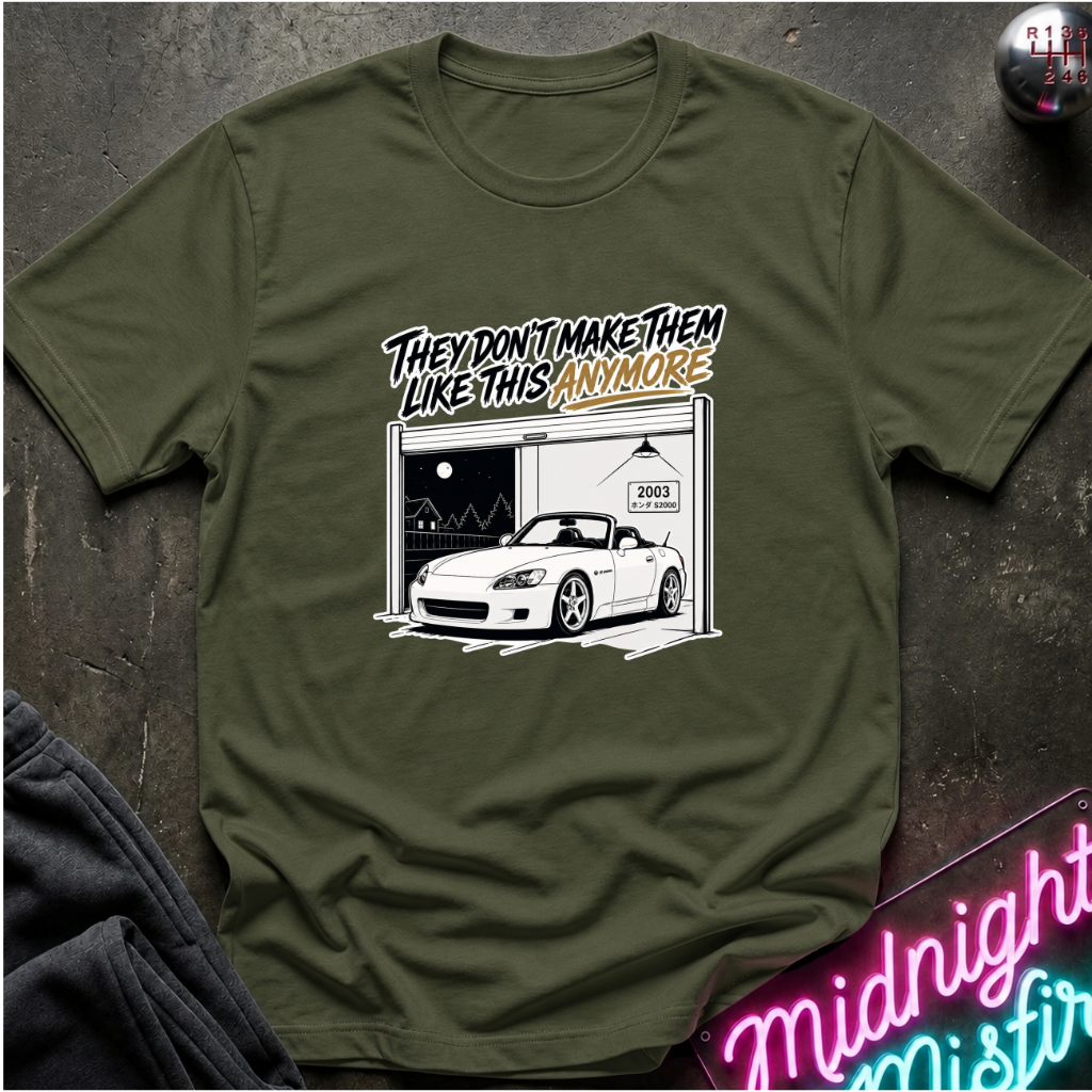 An olive green short-sleeve t-shirt laid flat on a dark textured surface. The shirt features a graphic print of a white convertible sports car parked in front of an open garage, with the text 'THEY DON'T MAKE THEM LIKE THIS ANYMORE' in stylized lettering.