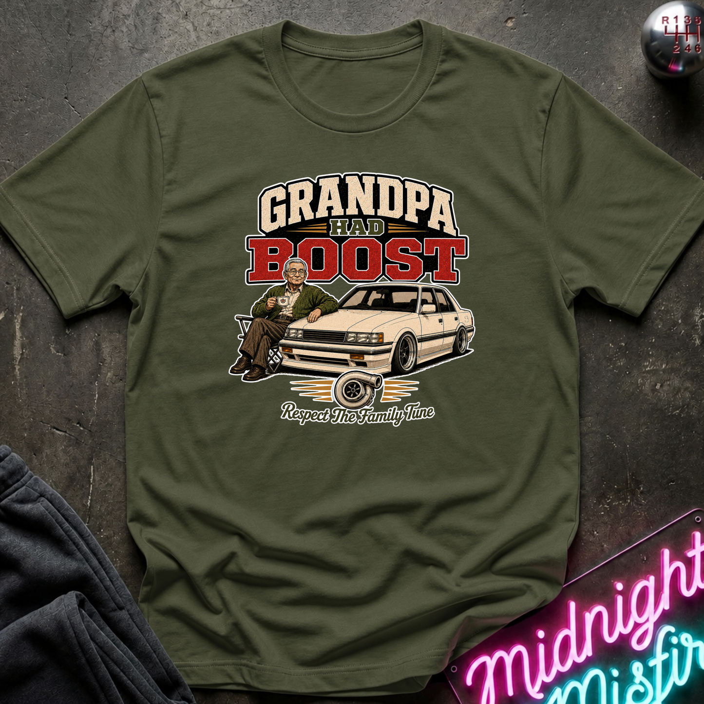 An olive green short-sleeve t-shirt with a vintage-style graphic that reads 'GRANDPA HAD BOOST' above an illustration of a man leaning on a car. The shirt is laid flat on a dark textured surface with a neon sign and dark fabric partially visible at the edges.