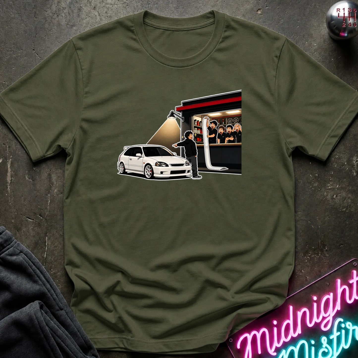 An olive green short-sleeve t-shirt laid flat on a dark textured surface, featuring a graphic print of a white hatchback car and a figure in front of a building. A neon sign reading 'Midnight Misfire' and folded dark fabric are partially visible around it.