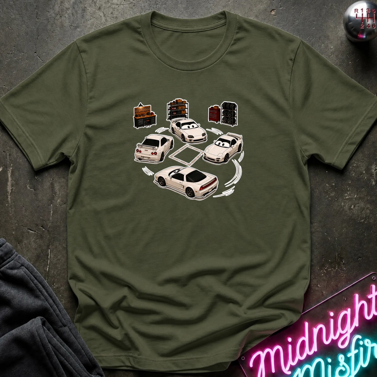 An olive green short-sleeve t-shirt laid flat on a dark textured surface, featuring a graphic print of several white sports cars arranged in a circle with drift-like tire marks.