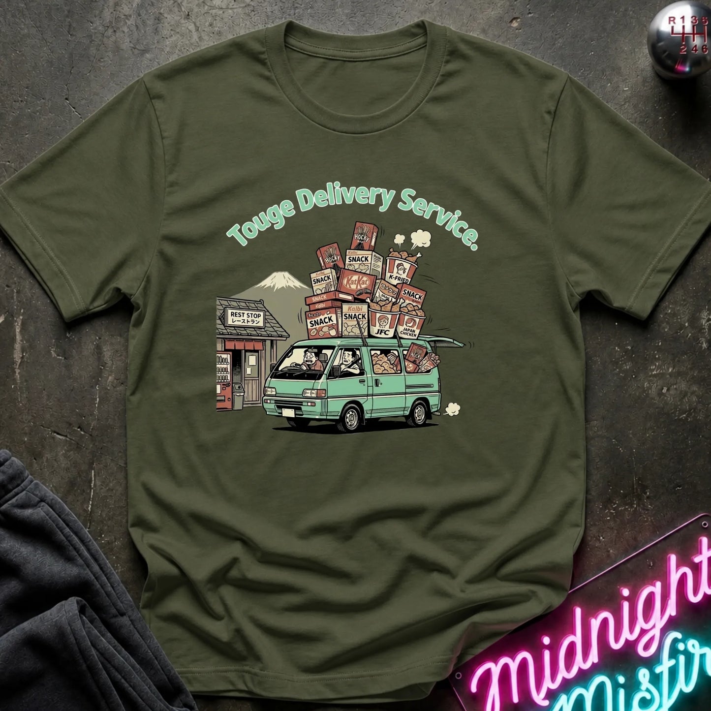 An olive green crewneck t-shirt featuring a retro graphic of a mint green van carrying stacks of ramen bowls under the text 'Touge Delivery Service.' The shirt is laid flat on a dark textured surface with a neon sign and dark fabric partially visible in the corners.