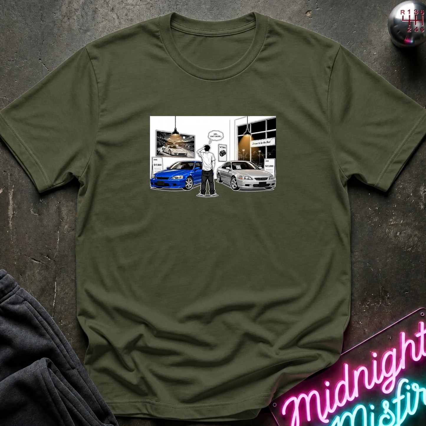 An olive green crewneck t-shirt laid flat on a dark textured surface, featuring a graphic print of a person standing between a blue car and a silver car in a garage.