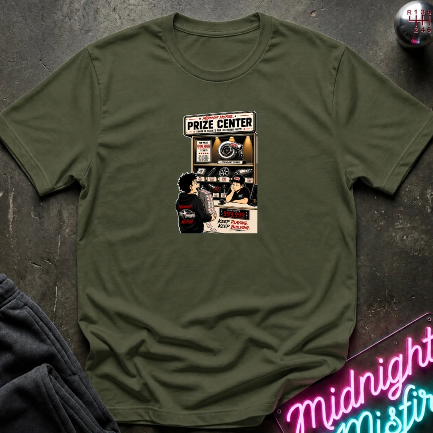 A olive green short-sleeve crewneck t-shirt laid flat on a dark textured surface, featuring a central graphic print of a prize center for car parts.