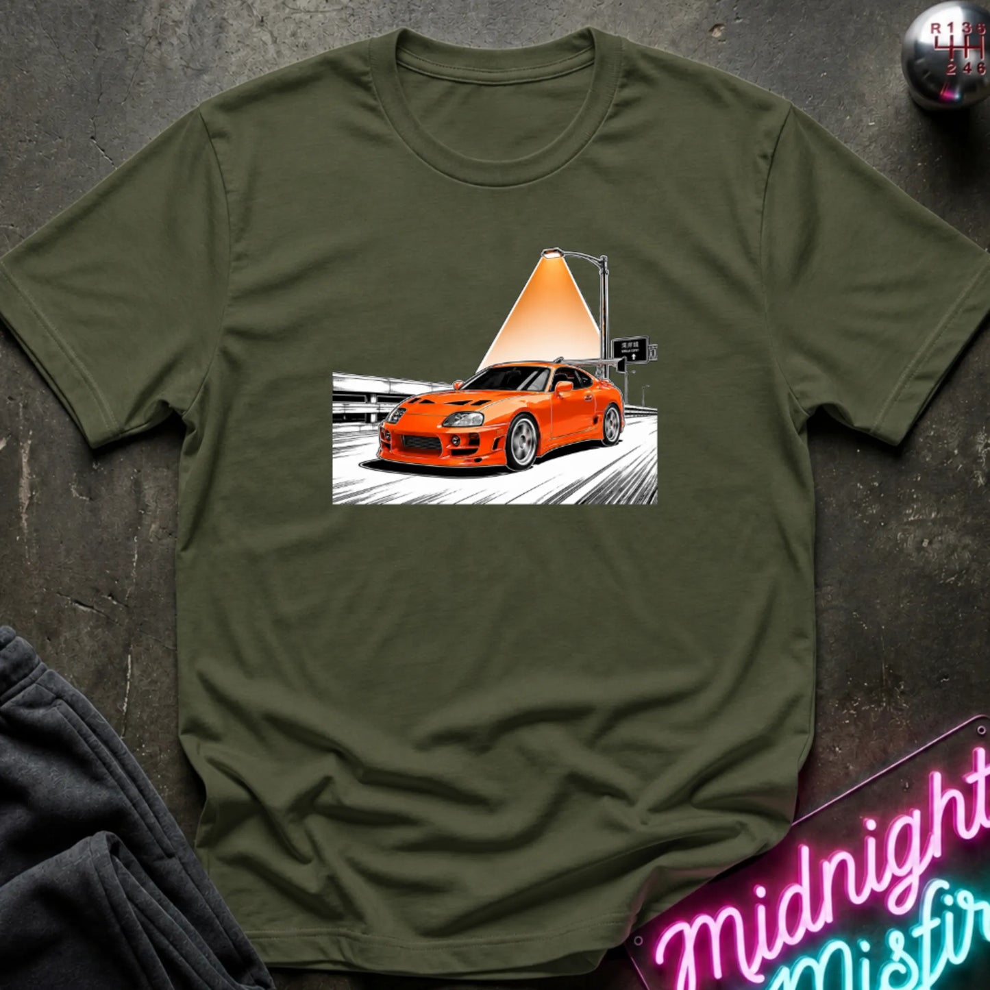 A olive green short-sleeve t-shirt laid flat on a dark textured surface, featuring a graphic print of an orange sports car speeding on highway.