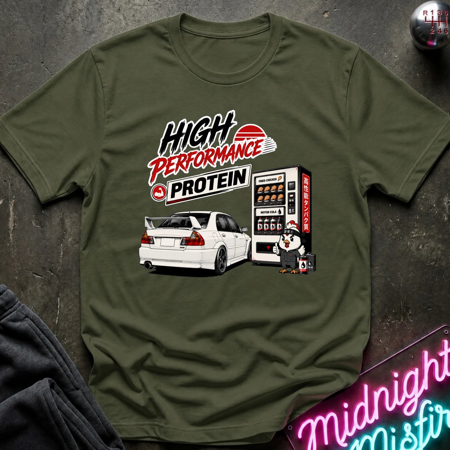 An olive green graphic t-shirt laid flat on a dark textured surface. The graphic features the text 'HIGH PERFORMANCE PROTEIN' above an illustration of a white sports car and a vending machine.