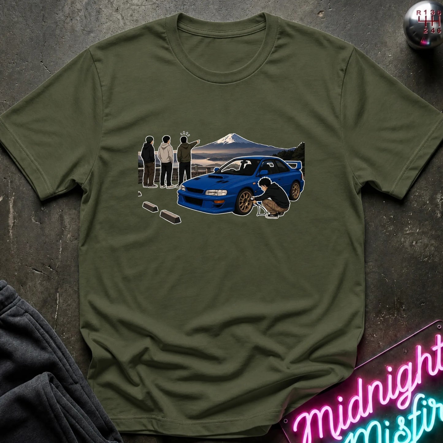 An olive green crewneck t-shirt featuring a graphic print of a blue sports car, a mountain backdrop, and several characters, laid flat on a dark textured surface with a neon sign and dark sweatpants partially visible.