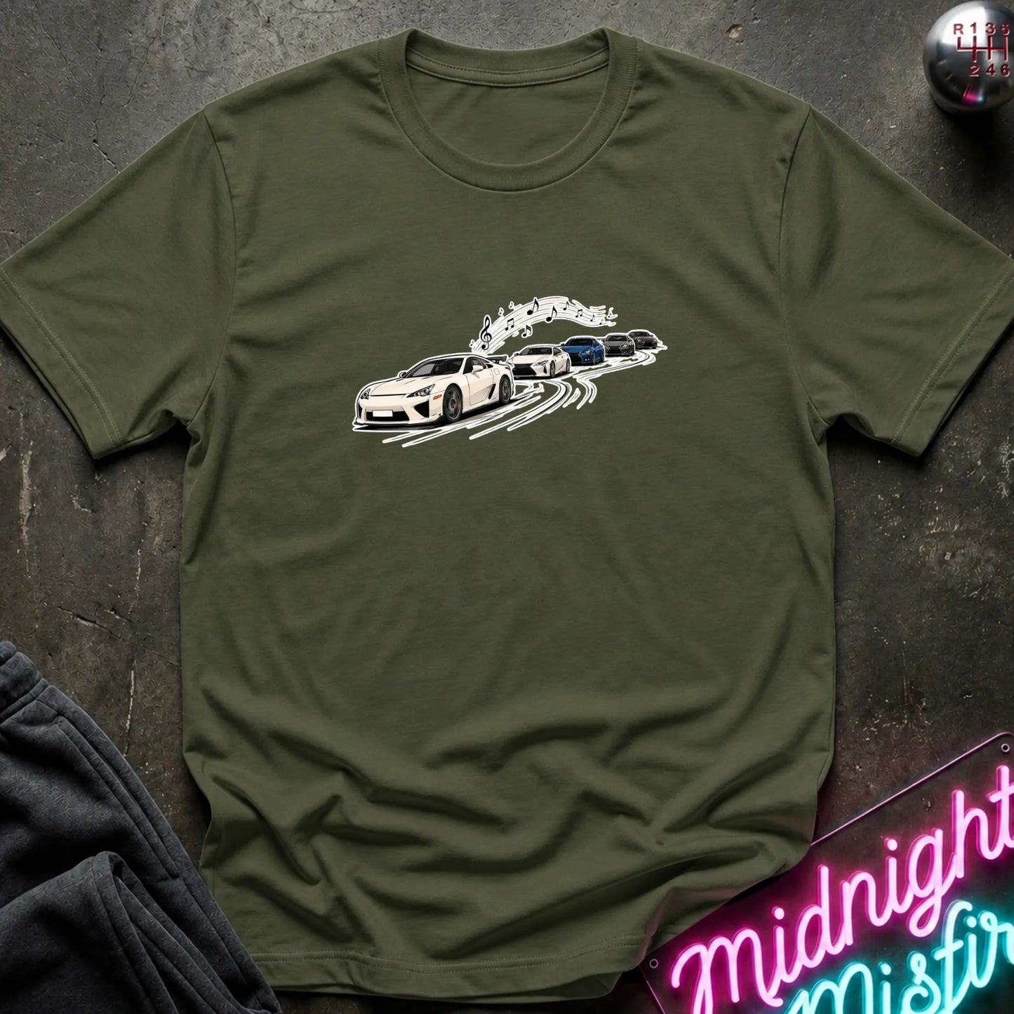 An olive green short-sleeve crewneck t-shirt featuring a graphic print of a white sports car leading a line of other cars, laid flat on a dark textured surface with a neon sign and dark pants partially visible.