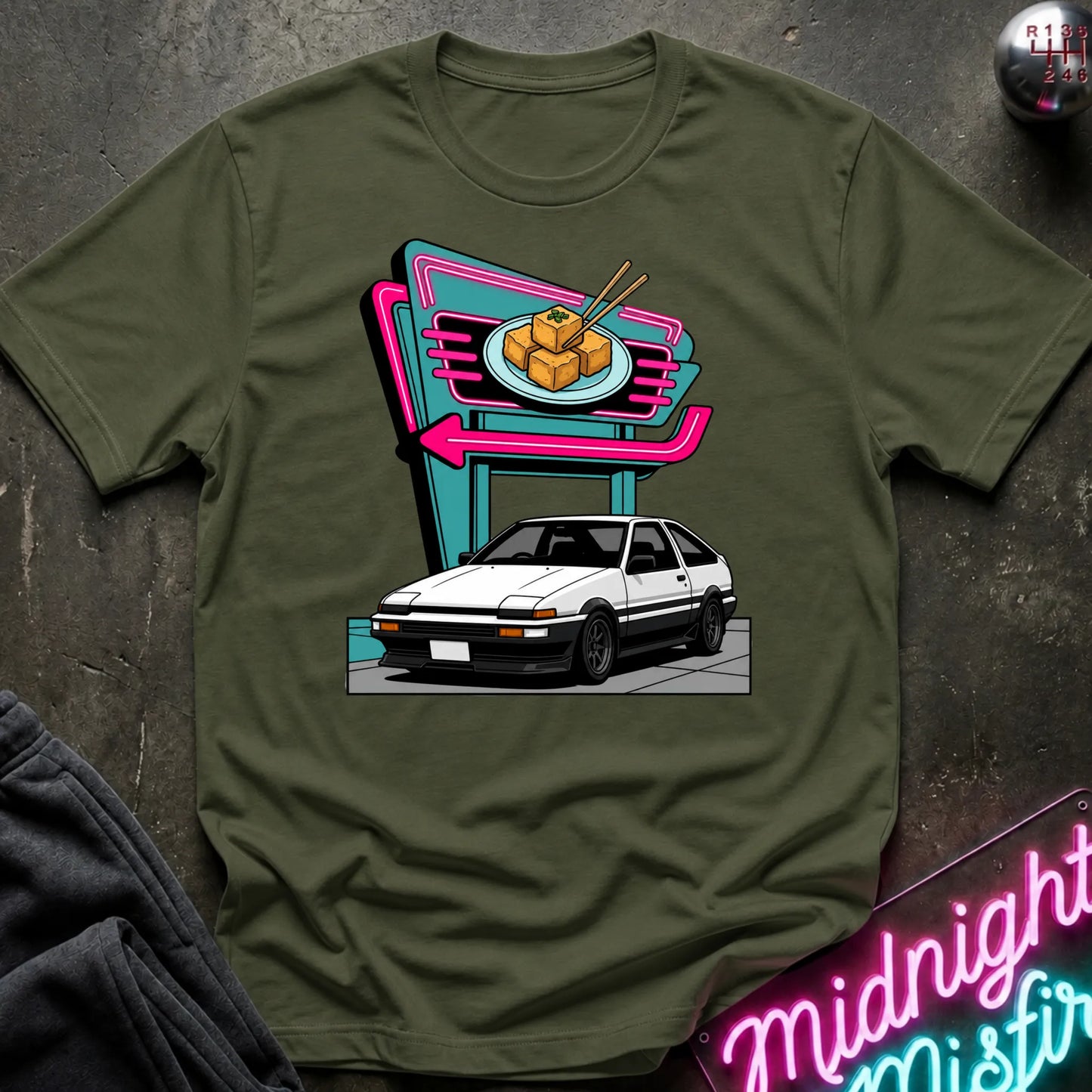 An olive green short-sleeve crewneck t-shirt featuring a retro-style graphic of a white sports car parked in front of a neon sign. The shirt is displayed flat on a dark textured surface with a dark sweatshirt and a neon sign partially visible as props.