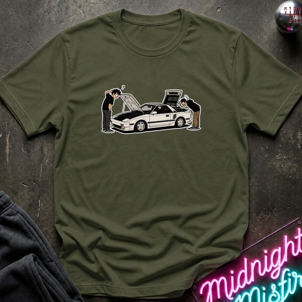 An olive green short-sleeve t-shirt with a graphic of a white sports car and two people, laid flat on a dark textured surface with a neon sign and dark pants in the background.