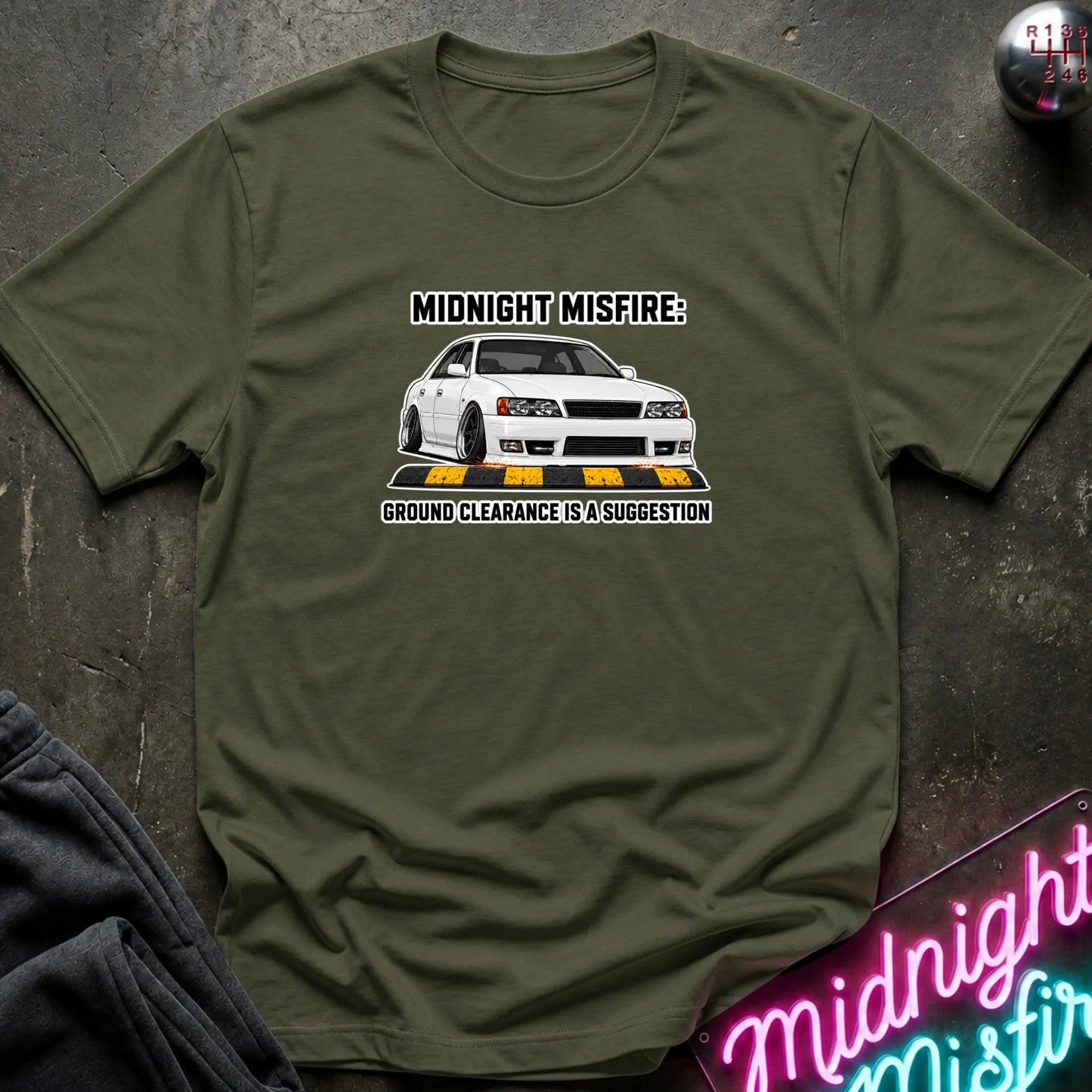 An olive green short-sleeve t-shirt with a graphic of a white Nissan Skyline R34 and the text 'MIDNIGHT MISFIRE: GROUND CLEARANCE IS A SUGGESTION'. The shirt is laid flat on a dark textured surface with a neon sign and dark pants partially visible.