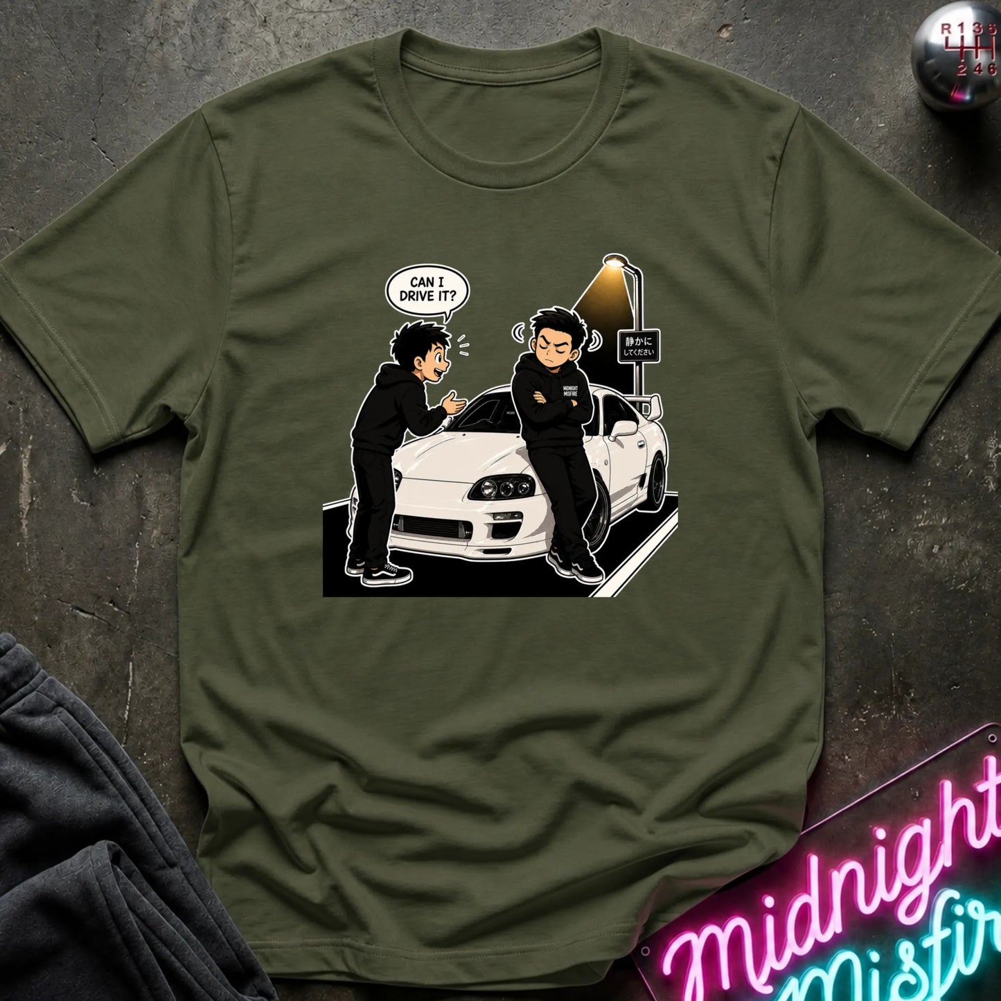 An olive green crewneck t-shirt featuring a cartoon graphic of two people and a white sports car, styled flat on a dark textured surface with a neon sign in the corner.