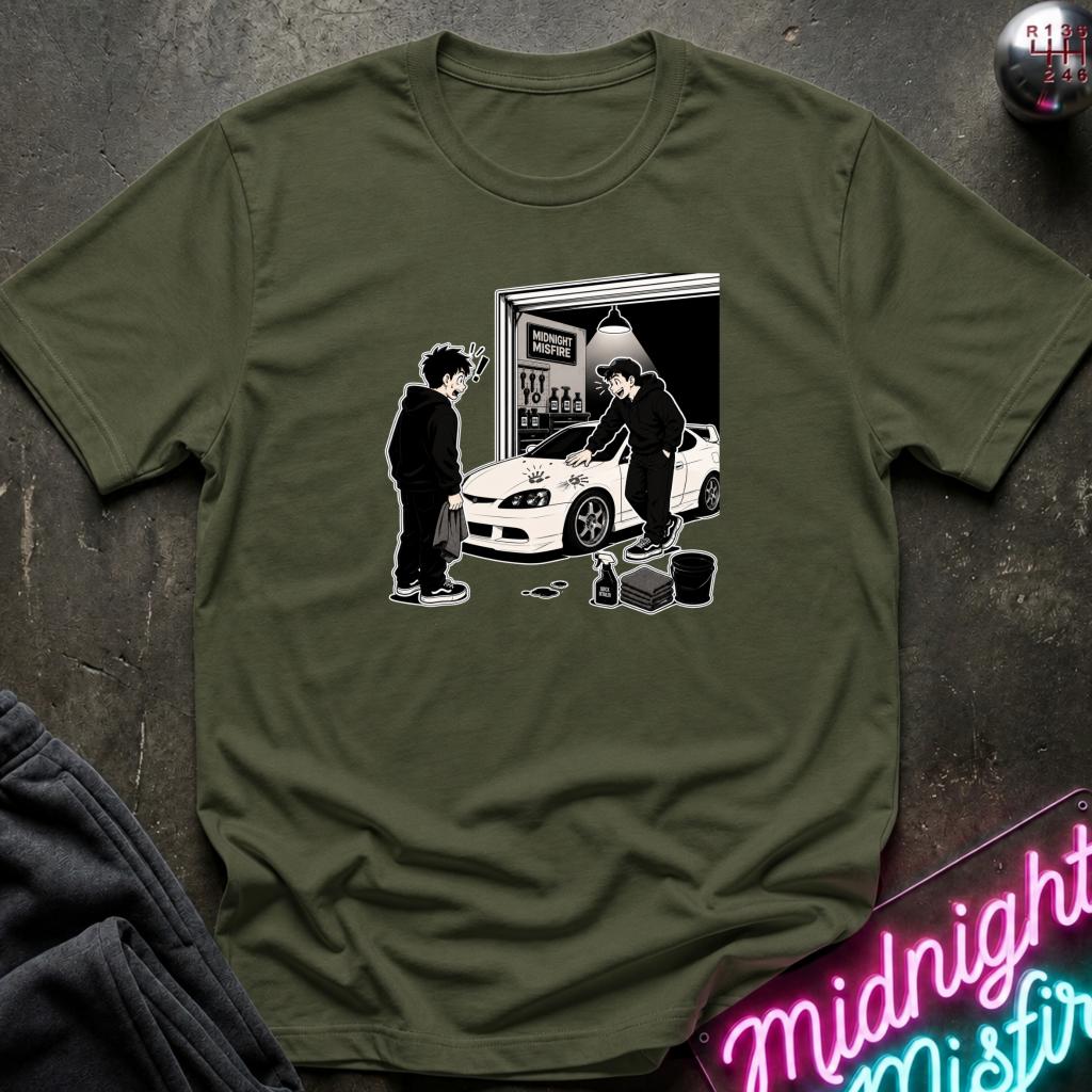 An olive green short-sleeve t-shirt laid flat on a dark textured surface, featuring a black and white graphic print of a person standing next to a white car in front of a garage door.