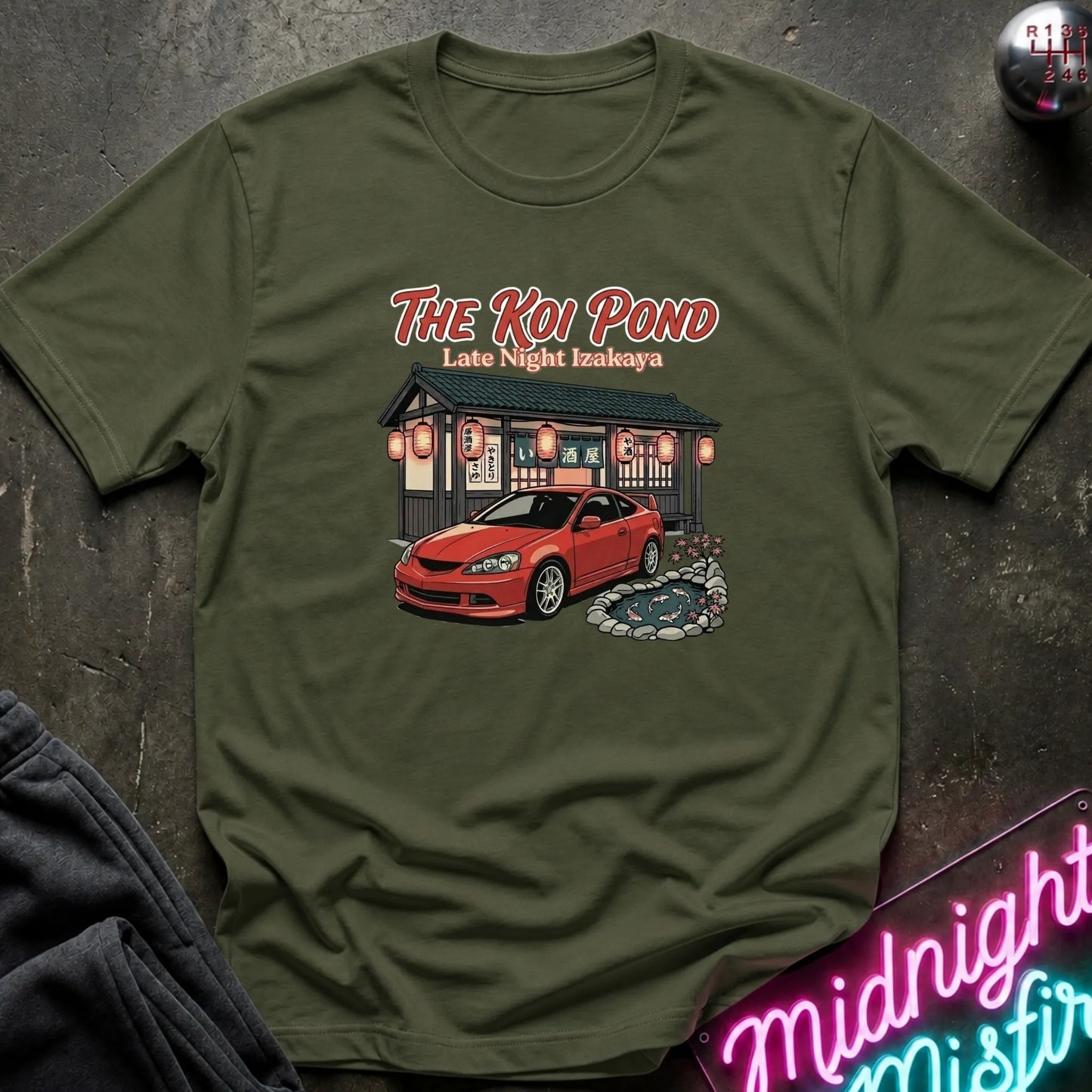 An olive green short-sleeve graphic t-shirt laid flat on a dark textured surface. The graphic features the text 'THE KOI POND Late Night Hamburger' above an illustration of a red sports car parked in front of a restaurant with red lanterns and a koi pond.