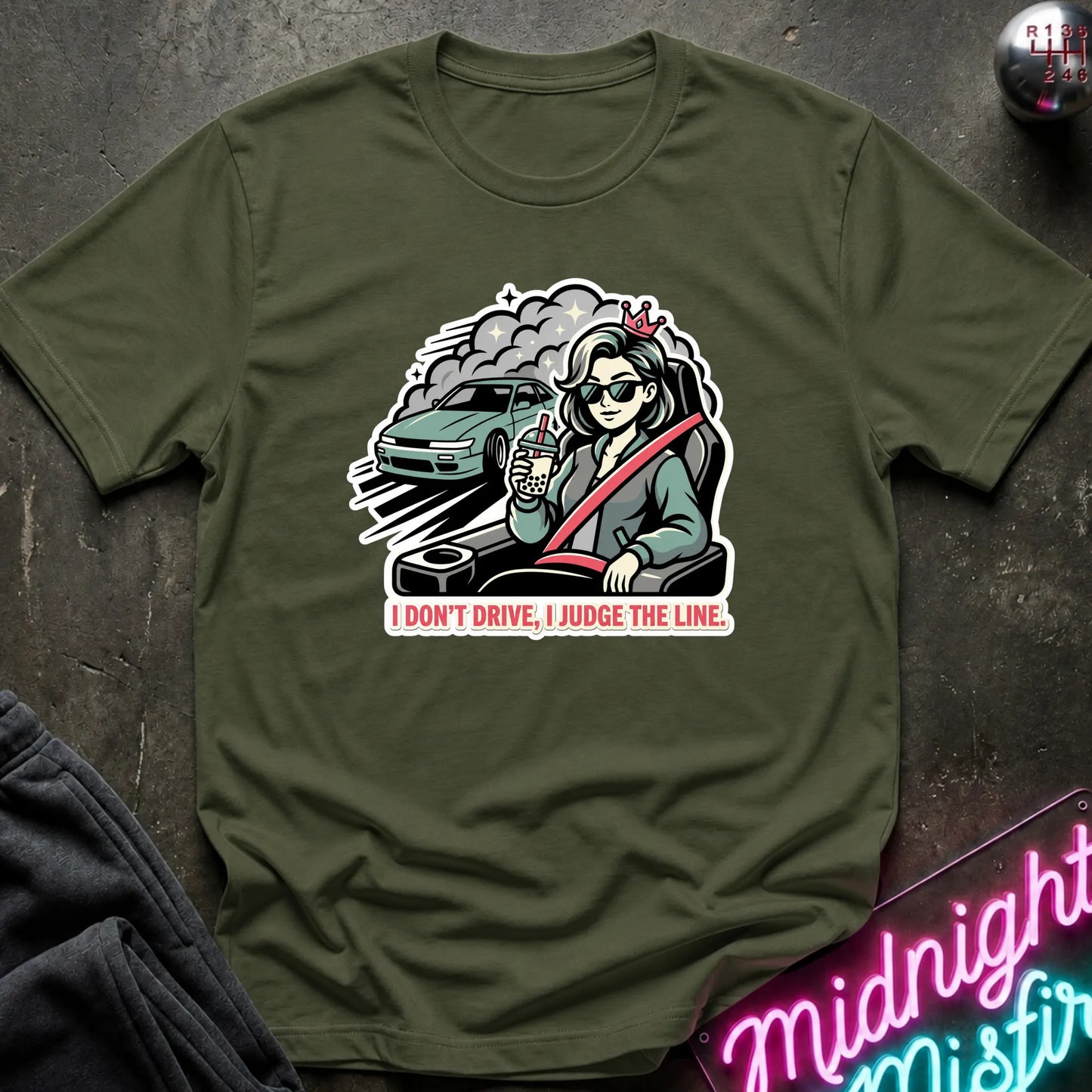 An olive green short-sleeve t-shirt laid flat on a dark textured surface, featuring a graphic of a woman in sunglasses driving a car with the text 'I DON'T DRIVE, I JUDGE THE LINE.'.