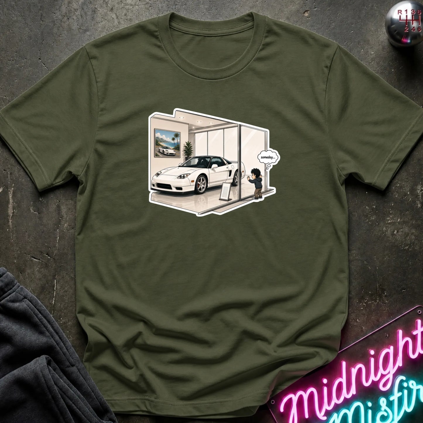 An olive green short-sleeve t-shirt laid flat on a dark textured surface, featuring a central graphic print of a white sports car inside a showroom with a cartoon character and a speech bubble.