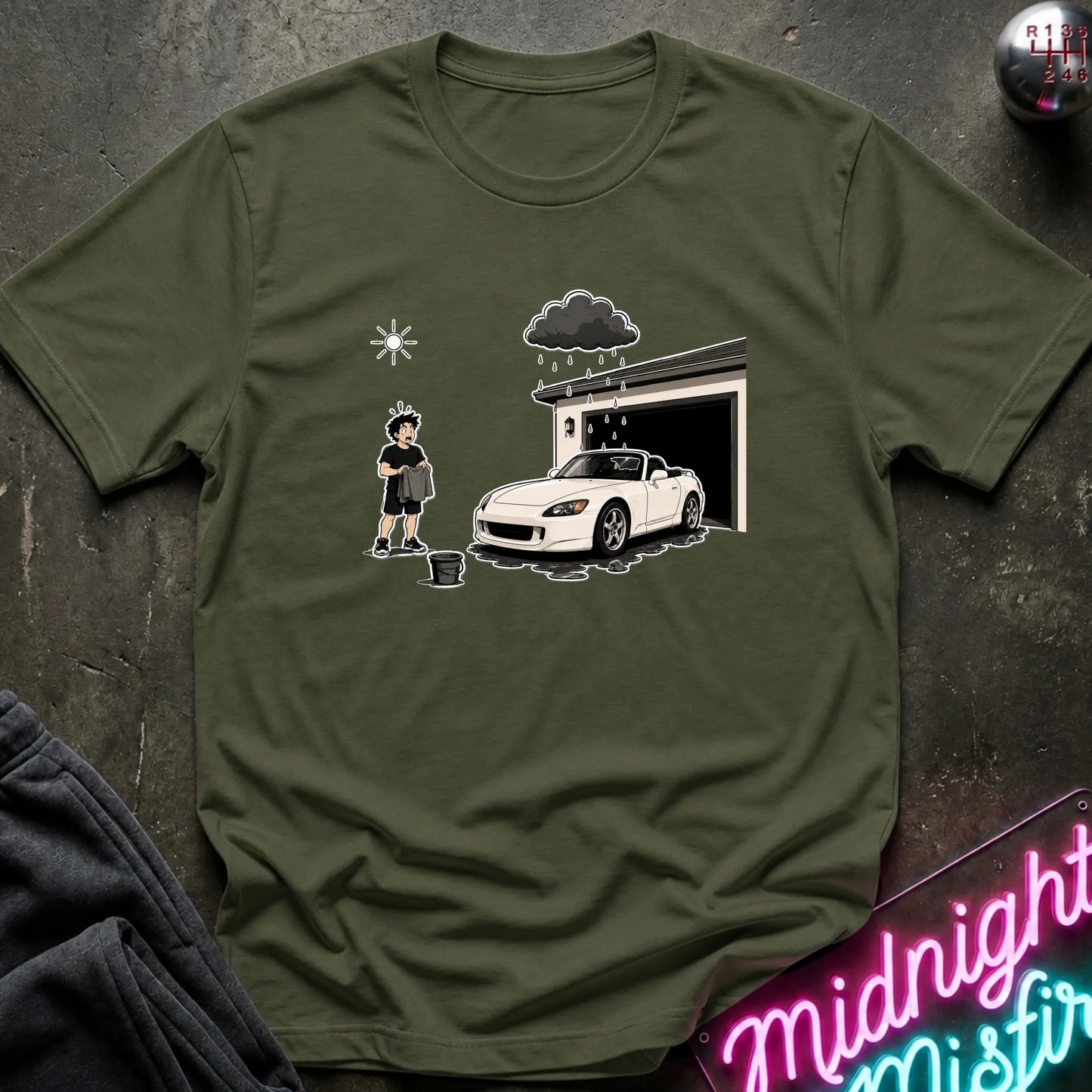 An olive green crewneck t-shirt featuring a cartoon graphic of a person washing a white sports car outside a garage under a dark raincloud, laid flat on a dark textured surface.