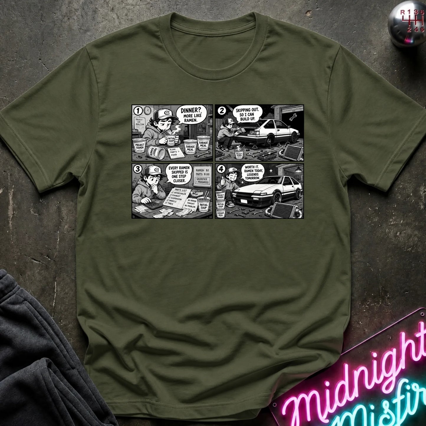 An olive green crewneck t-shirt featuring a black and white comic strip graphic of a police officer and a car, laid flat on a dark textured surface with a neon sign and dark fabric in the background.