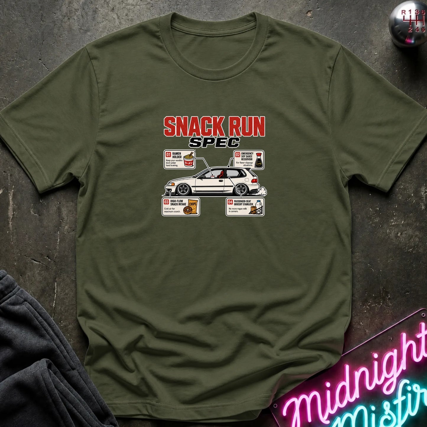 An olive green short-sleeve t-shirt with a 'SNACK RUN SPEC' graphic on the chest, laid flat on a dark textured surface with a neon sign and dark fabric in the corners.