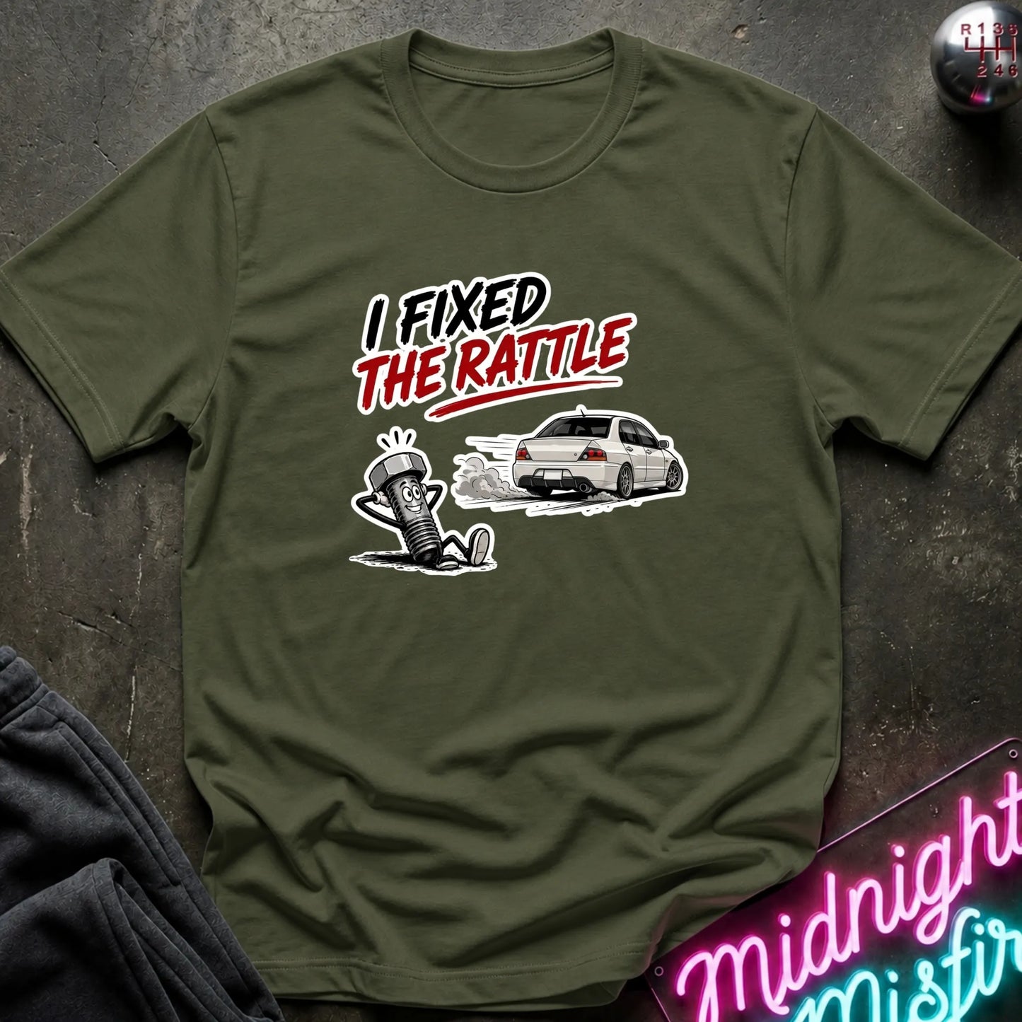 An olive green graphic t-shirt laid flat on a dark textured surface. The graphic features the text 'I FIXED THE RATTLE' in black and red, above a cartoon character and a white car.