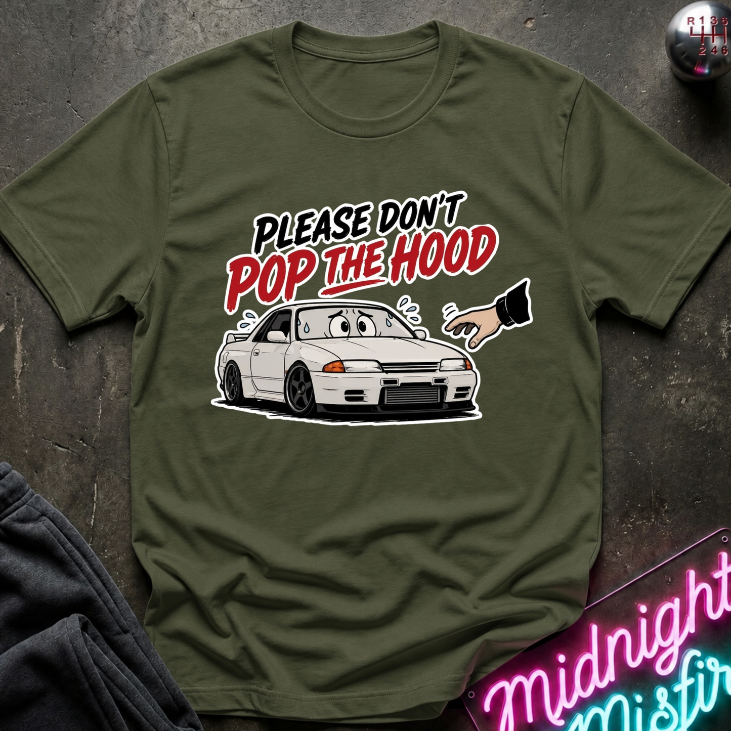 An olive green graphic t-shirt laid flat on a dark textured surface. The shirt features a cartoon illustration of a white sports car with a surprised face, alongside a hand pointing, under the text 'PLEASE DON'T POP THE HOOD' in black, white, and red lettering.
