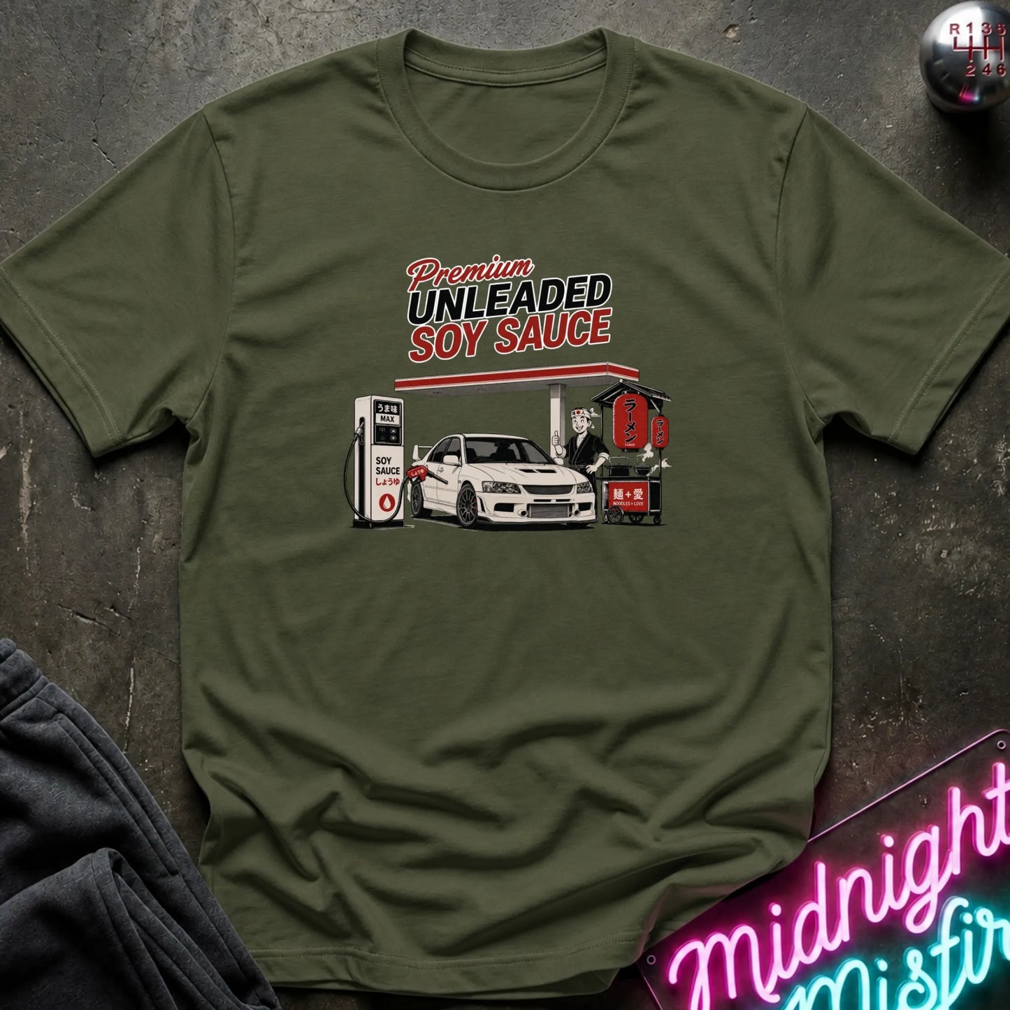 An olive green short-sleeve t-shirt with a retro-style graphic of a gas station attendant pumping fuel next to a white car under the text 'Premium UNLEADED SOY SAUCE'. The shirt is laid flat on a dark textured surface with a neon sign and dark fabric partially visible in the corners.
