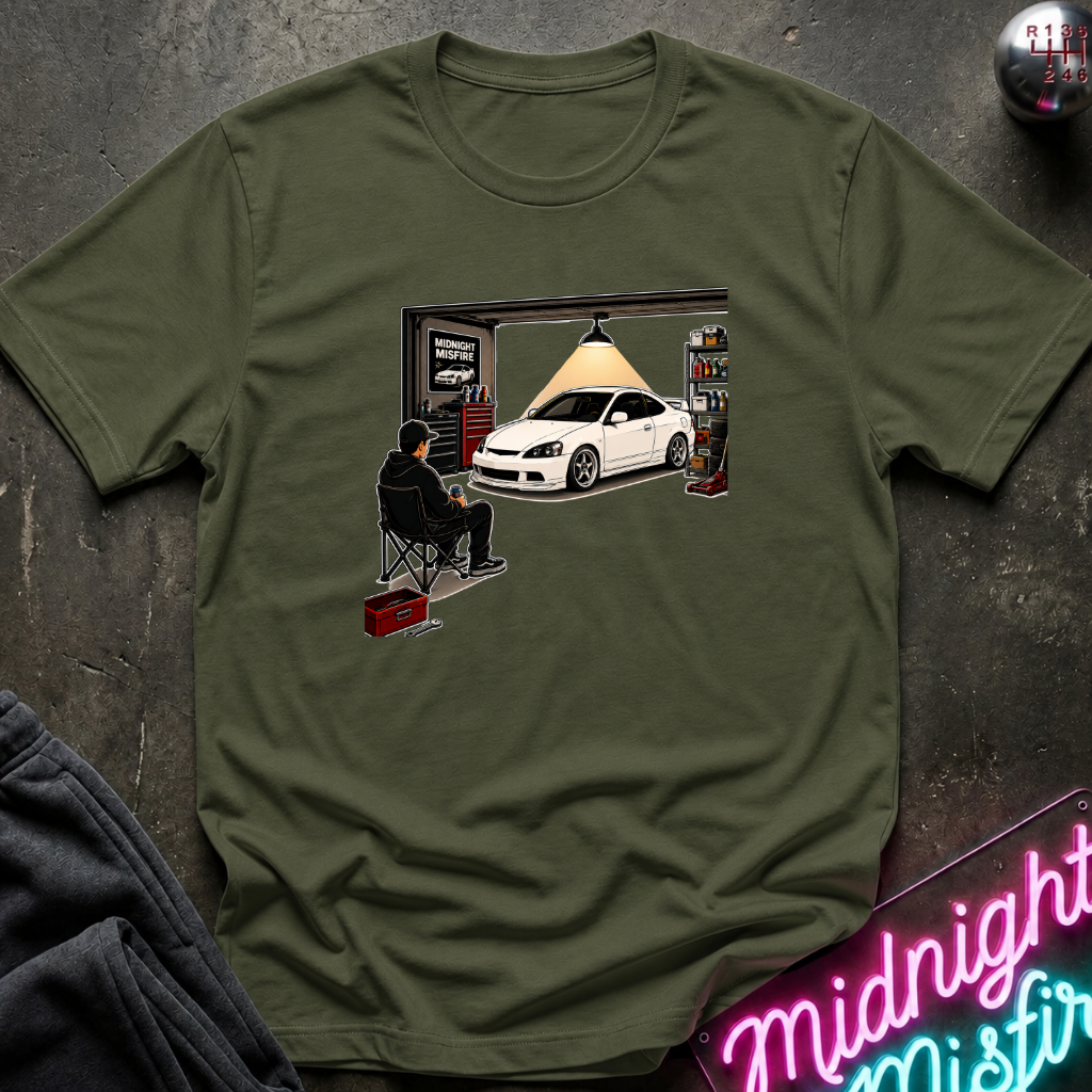 An olive green short-sleeve t-shirt with a graphic print of a white sports car in a garage scene, styled flat on a dark textured background with a neon sign and dark jacket partially visible.