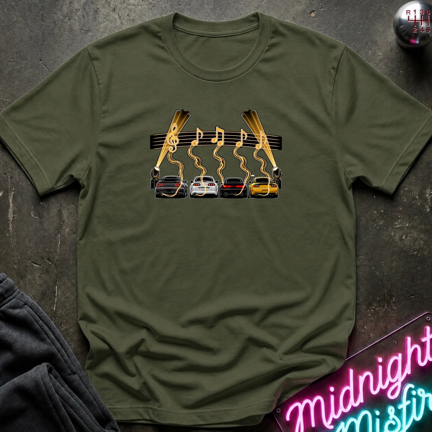 An olive green short-sleeve t-shirt laid flat on a dark textured surface, featuring a graphic print of four cars (red, white, gray, yellow) with gold wavy lines and musical notes above them, flanked by two lights.