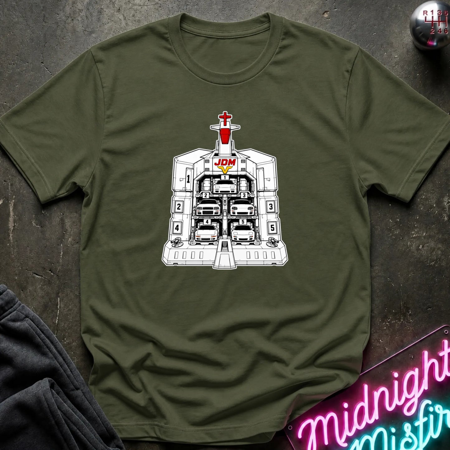 An olive green short-sleeve crewneck t-shirt laid flat on a dark textured surface, featuring a black and white graphic of the JDM (Japanese Domestic Market) cars in a launch bay.