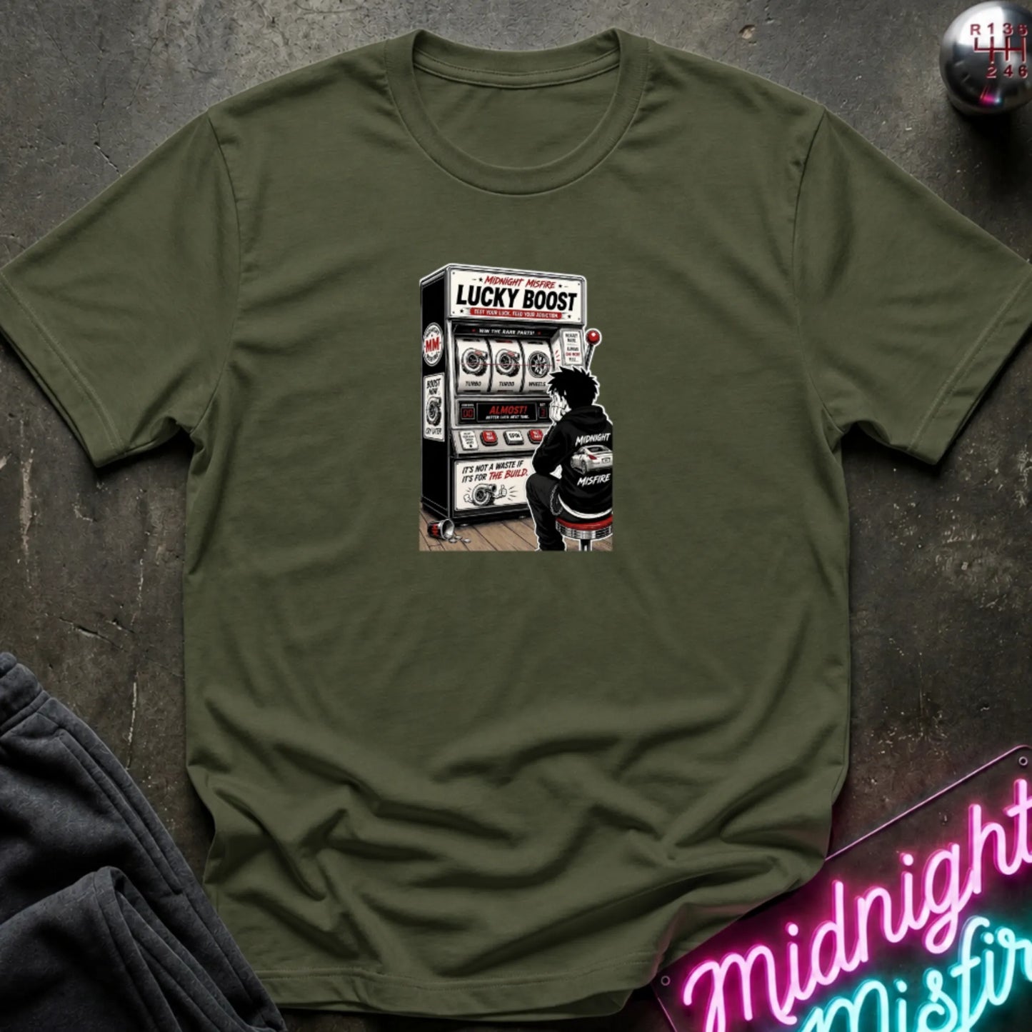 A olive green short-sleeve t-shirt with a retro-style graphic of a slot machine, displayed flat on a dark textured surface with a neon sign and sweatpants partially visible.