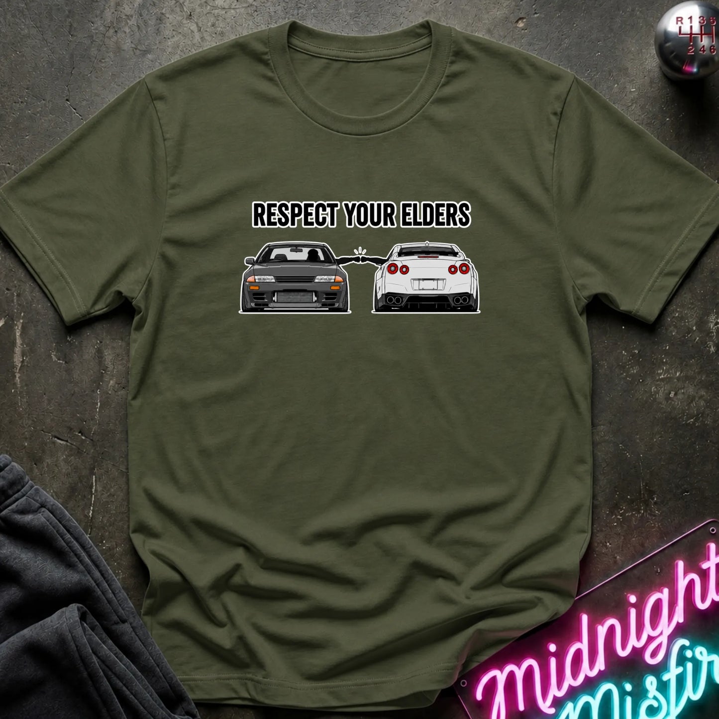 An olive green short-sleeve graphic T-shirt laid flat on a dark textured surface. The shirt has a black printed slogan reading 'RESPECT YOUR ELDERS' above two cars, with a neon sign and dark clothing partially visible around it.
