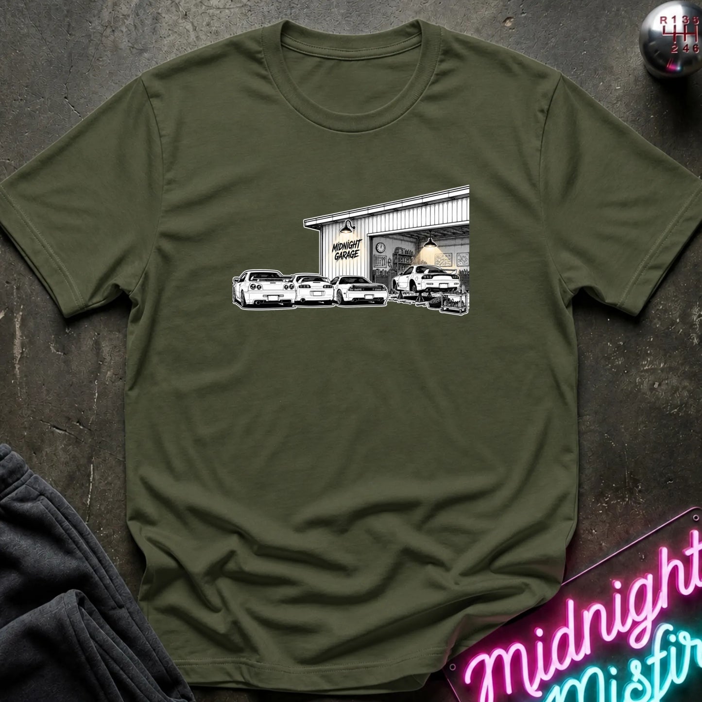 An olive green short-sleeve t-shirt laid flat on a dark textured surface, featuring a black and white graphic print of a garage with several cars in front.