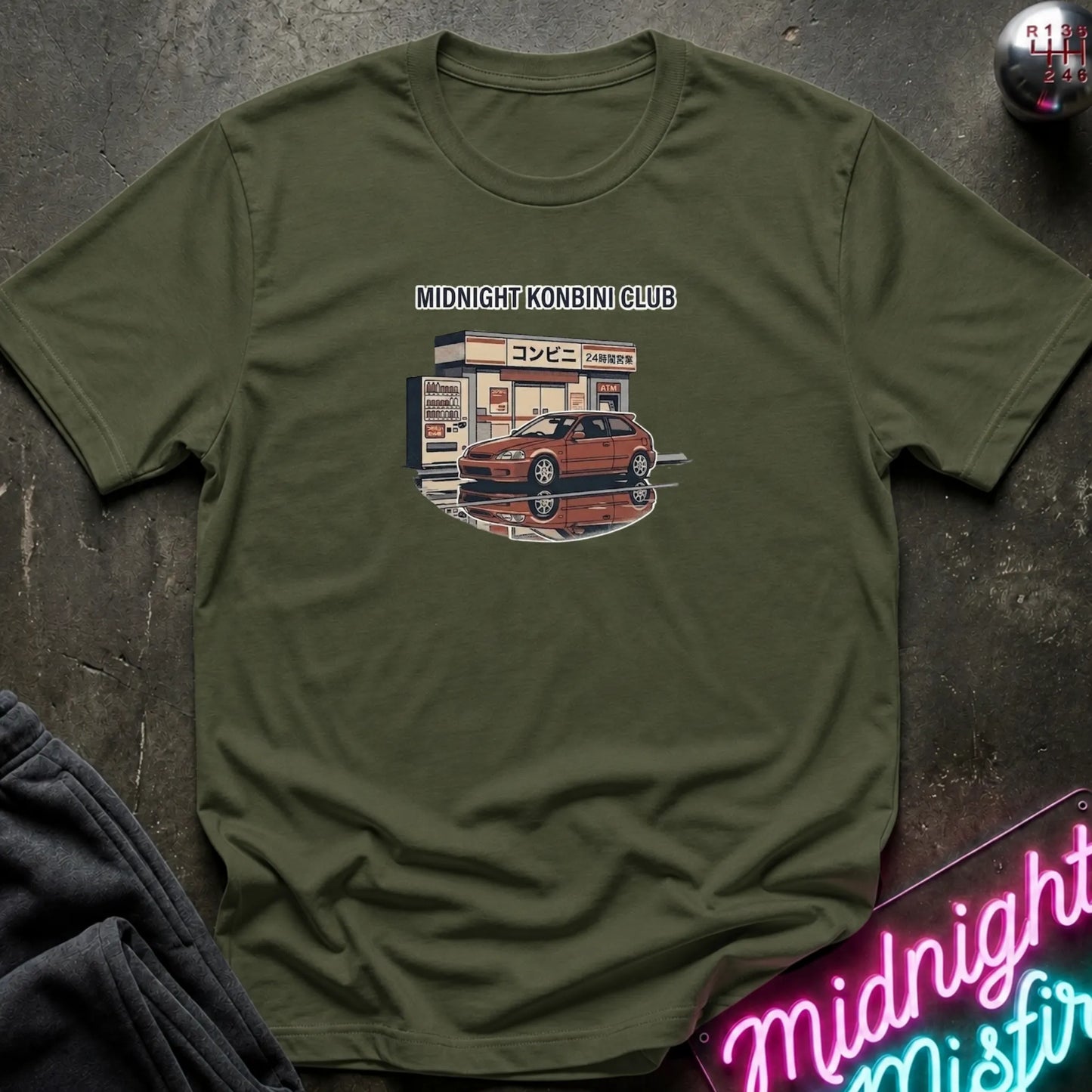 An olive green short-sleeve t-shirt with a graphic print of a red sports car in front of a convenience store under the text 'MIDNIGHT KONBINI CLUB'. The shirt is displayed flat on a dark textured surface with a neon sign and dark pants partially visible.