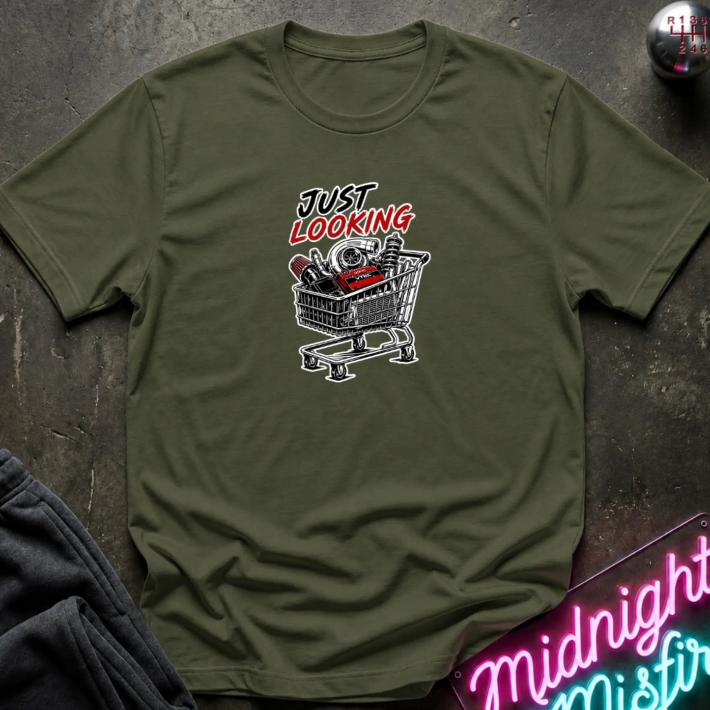 An olive green short-sleeve t-shirt with a graphic print that reads 'JUST LOOKING' above a shopping cart filled with items, laid flat on a dark textured surface with decorative props.