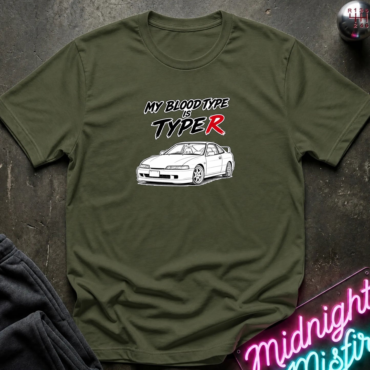 An olive green crewneck t-shirt laid flat on a dark textured surface. The shirt features a graphic print of a white sports car with the text 'MY BLOODTYPE IS TYPE R' in black and red lettering.