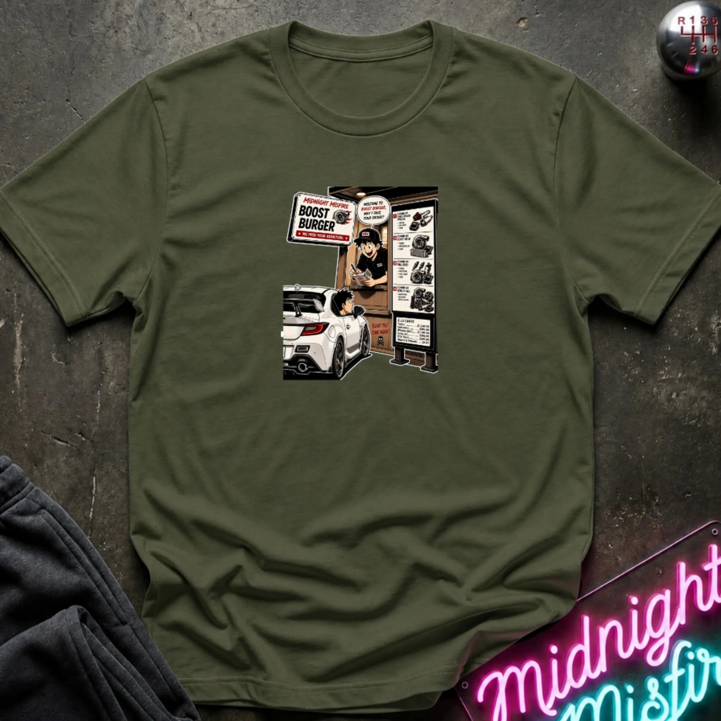 A olive green short-sleeve crewneck t-shirt laid flat on a dark textured surface, featuring a central graphic print of a street-food burger scene with a white car and signs.