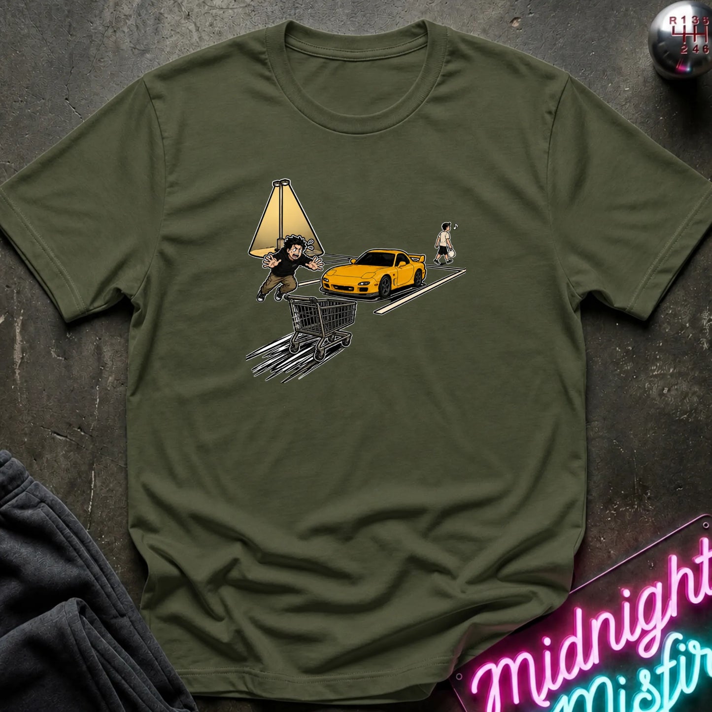 An olive green short-sleeve t-shirt laid flat on a dark textured surface, featuring a graphic print of a yellow sports car, a shopping cart, a lamp, and a small figure.
