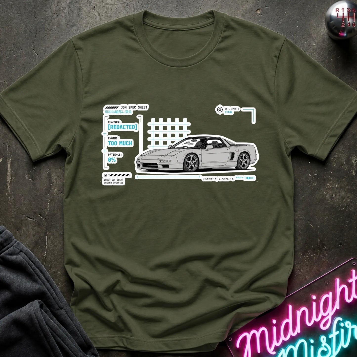 An olive green short-sleeve t-shirt laid flat on a dark textured surface, featuring a white and light blue graphic print of a sports car with technical-style labels. A neon sign and folded dark fabric are partially visible around the edges.