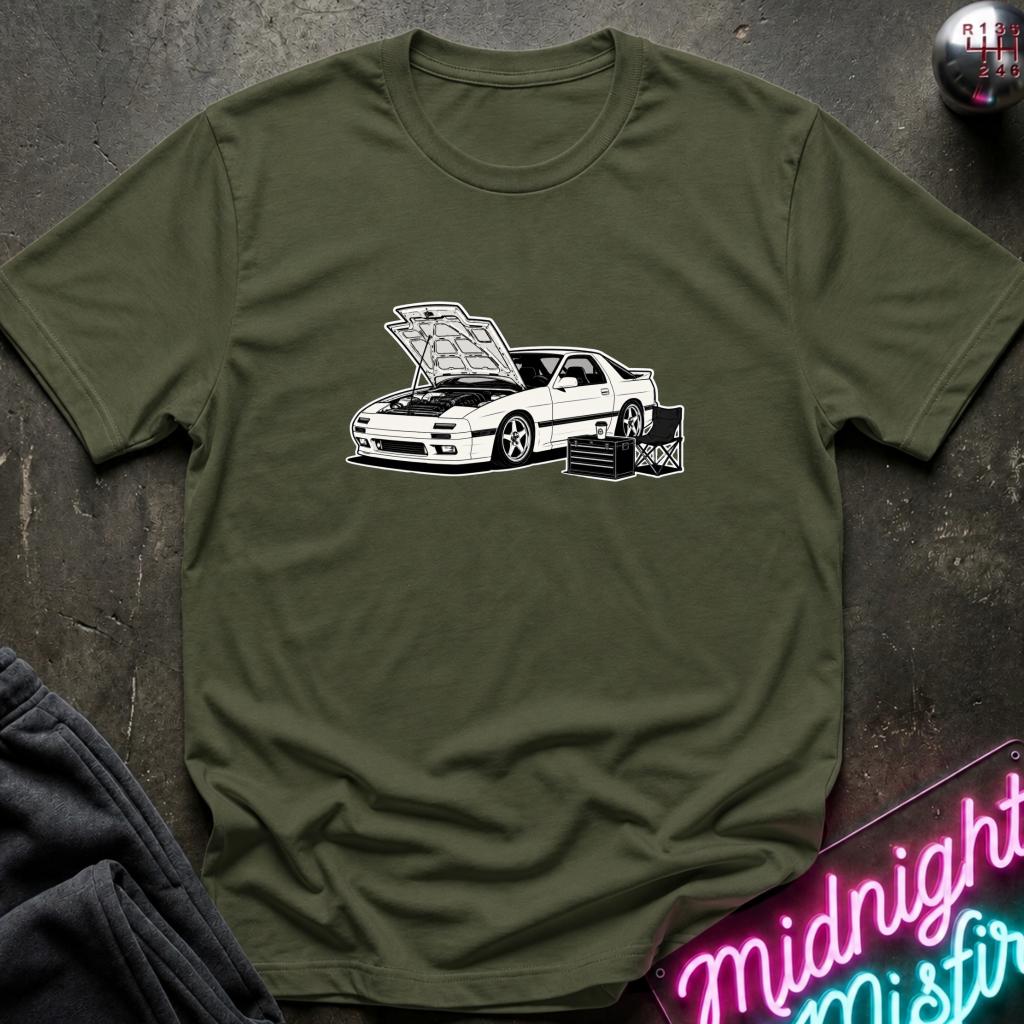 An olive green short-sleeve t-shirt with a white graphic of a sports car with its hood open, styled flat on a dark textured background with a neon sign and dark pants partially visible.